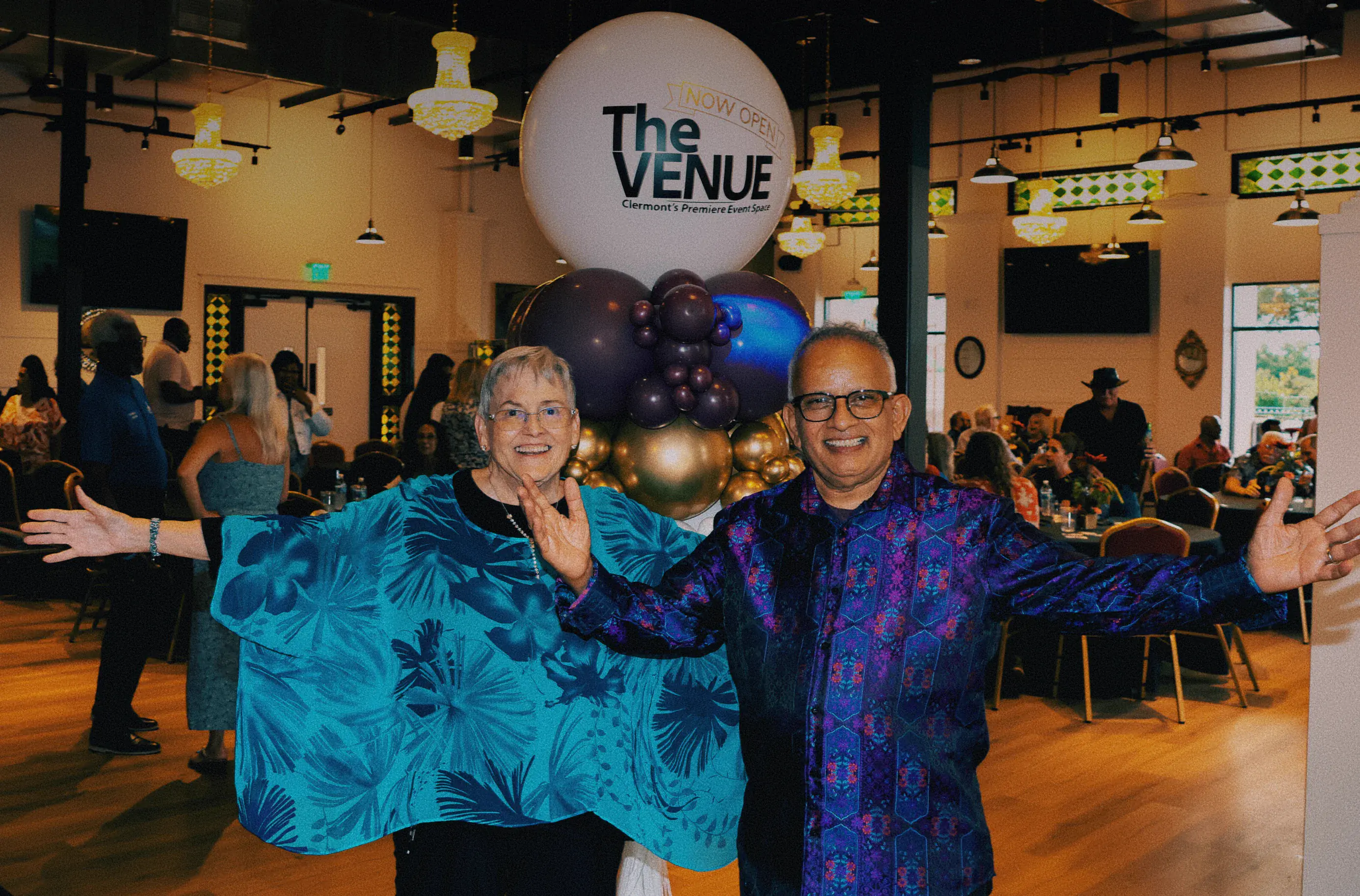 Uday Kadam and guest with arms open celebrating the grand opening of The Venue Clermont in front of a large NOW OPEN balloon display inside the festively decorated venue